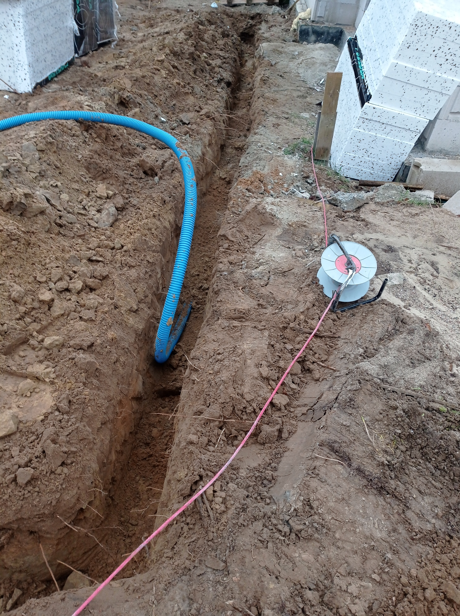 Wykop w ziemi z niebieską rurą karbowaną i szpulą z kablem. Przygotowanie do instalacji elektrycznej w gruncie.