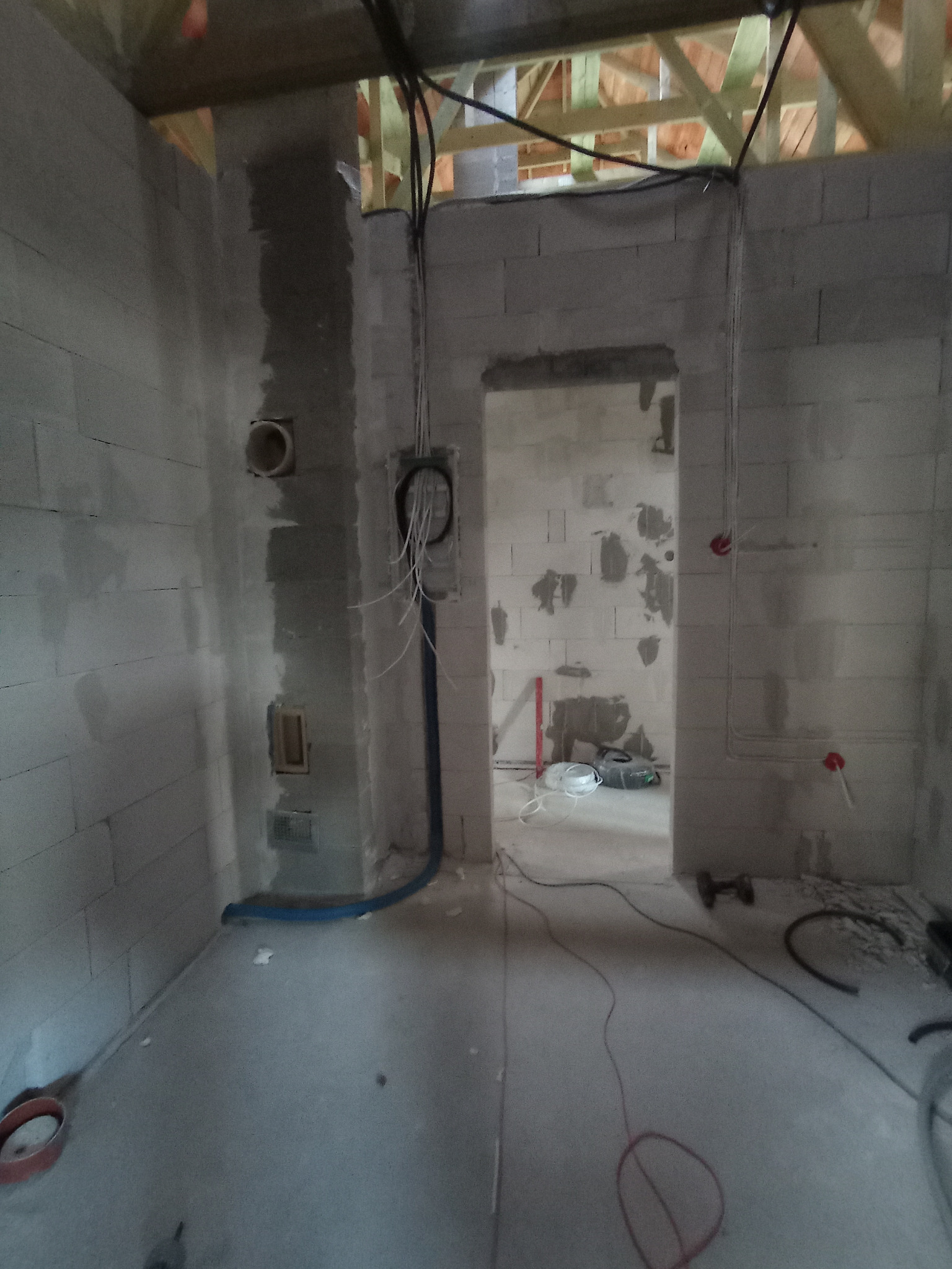 Surowy stan wnętrza z widoczną instalacją elektryczną w ścianach z betonu komórkowego. Kable i rury prowadzone pod stropem z drewnianą konstrukcją. W tle widoczne kolejne pomieszczenie.