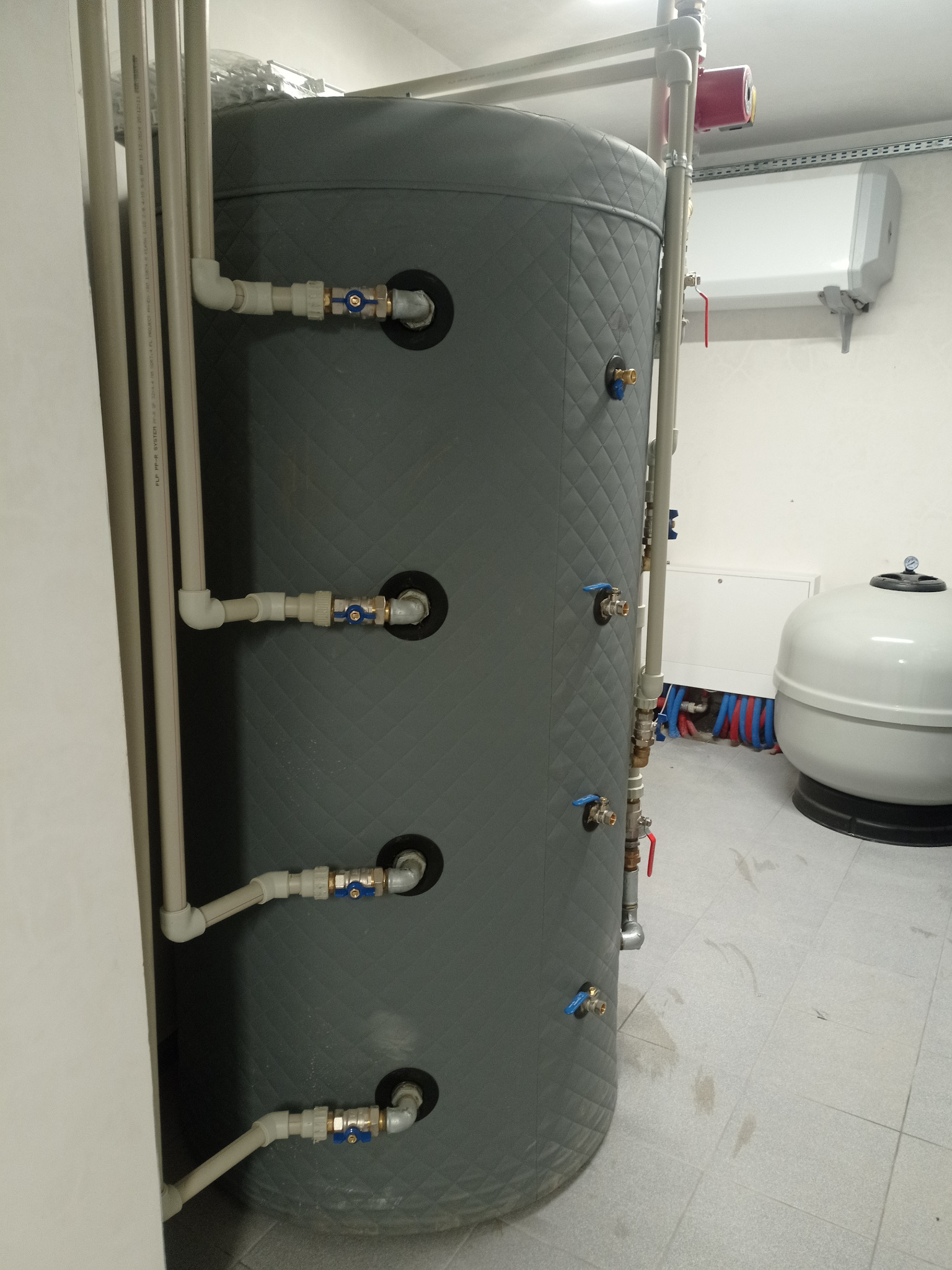 Szary, ocieplony zbiornik akumulacyjny z licznymi przyłączami rur i zaworami w kotłowni. Widoczne elementy instalacji grzewczej i podłączenia do ogrzewania podłogowego.