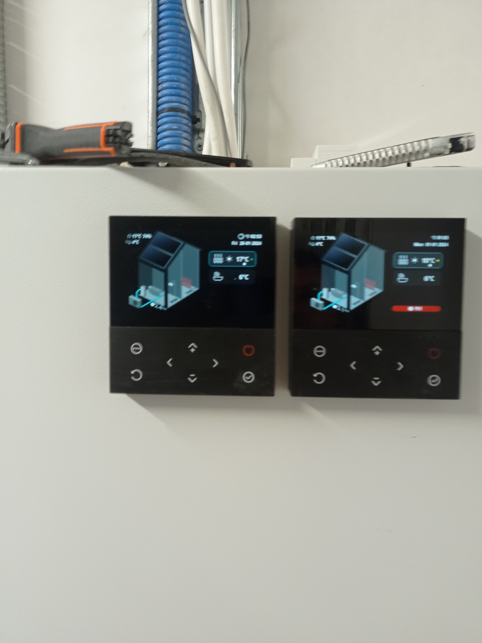 Dwa panele sterowania ogrzewaniem podłogowym z wyświetlaczami pokazującymi schemat instalacji i temperaturę. Nad panelami widoczne kable i narzędzia.