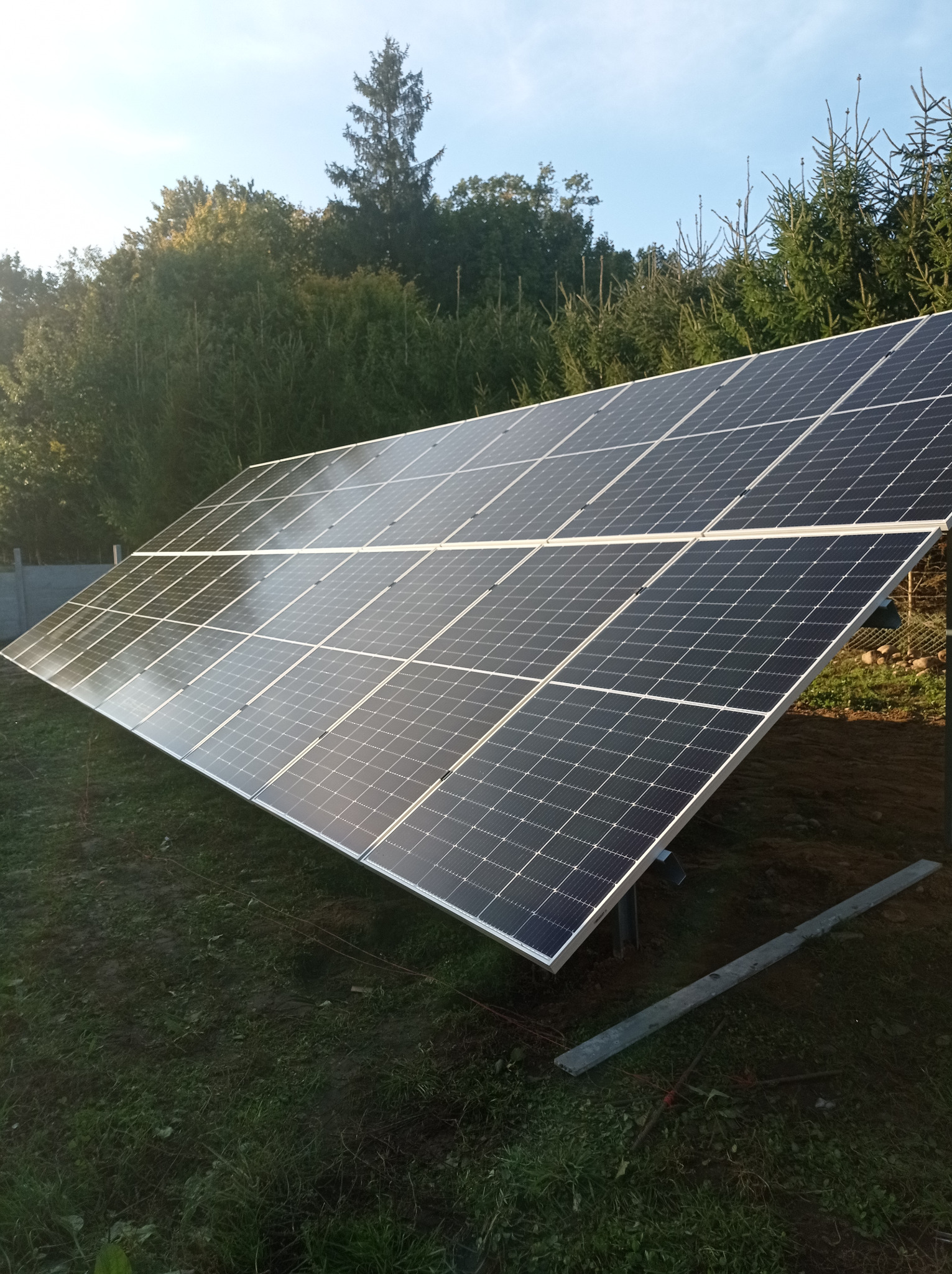 Panel słoneczny na tle zieleni, z widocznymi drzewami i błękitnym niebem w tle. Instalacja fotowoltaiczna na trawiastym terenie, w słoneczny dzień.