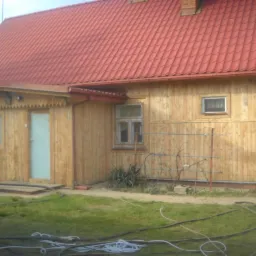 Drewniany dom z czerwonym dachem po renowacji. Świeżo pomalowane deski elewacyjne z widocznymi słojami. Na pierwszym planie trawa i węże.