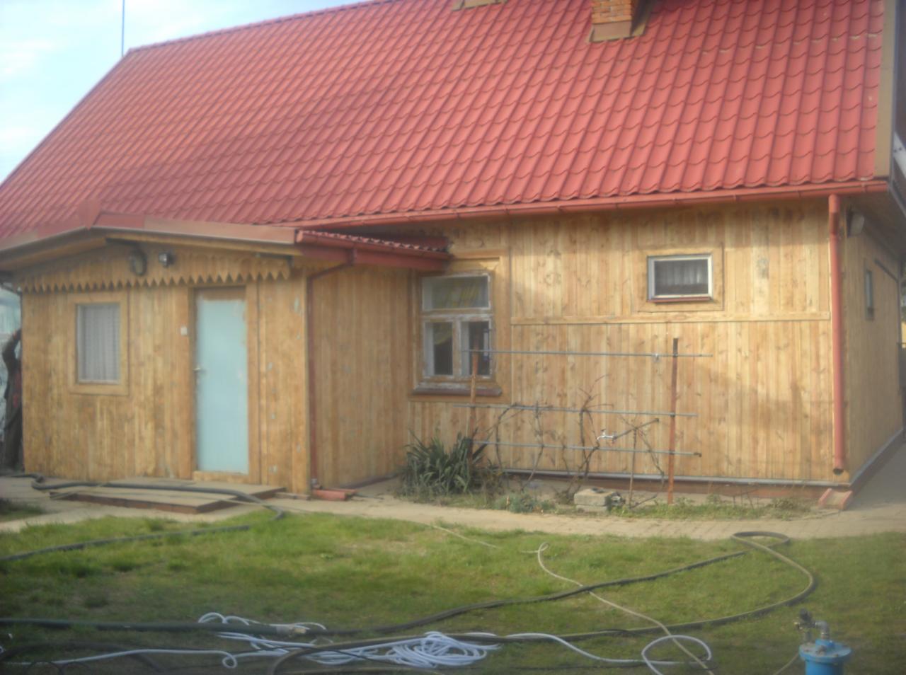 Drewniany dom z czerwonym dachem po renowacji. Świeżo pomalowane deski elewacyjne z widocznymi słojami. Na pierwszym planie trawa i węże.