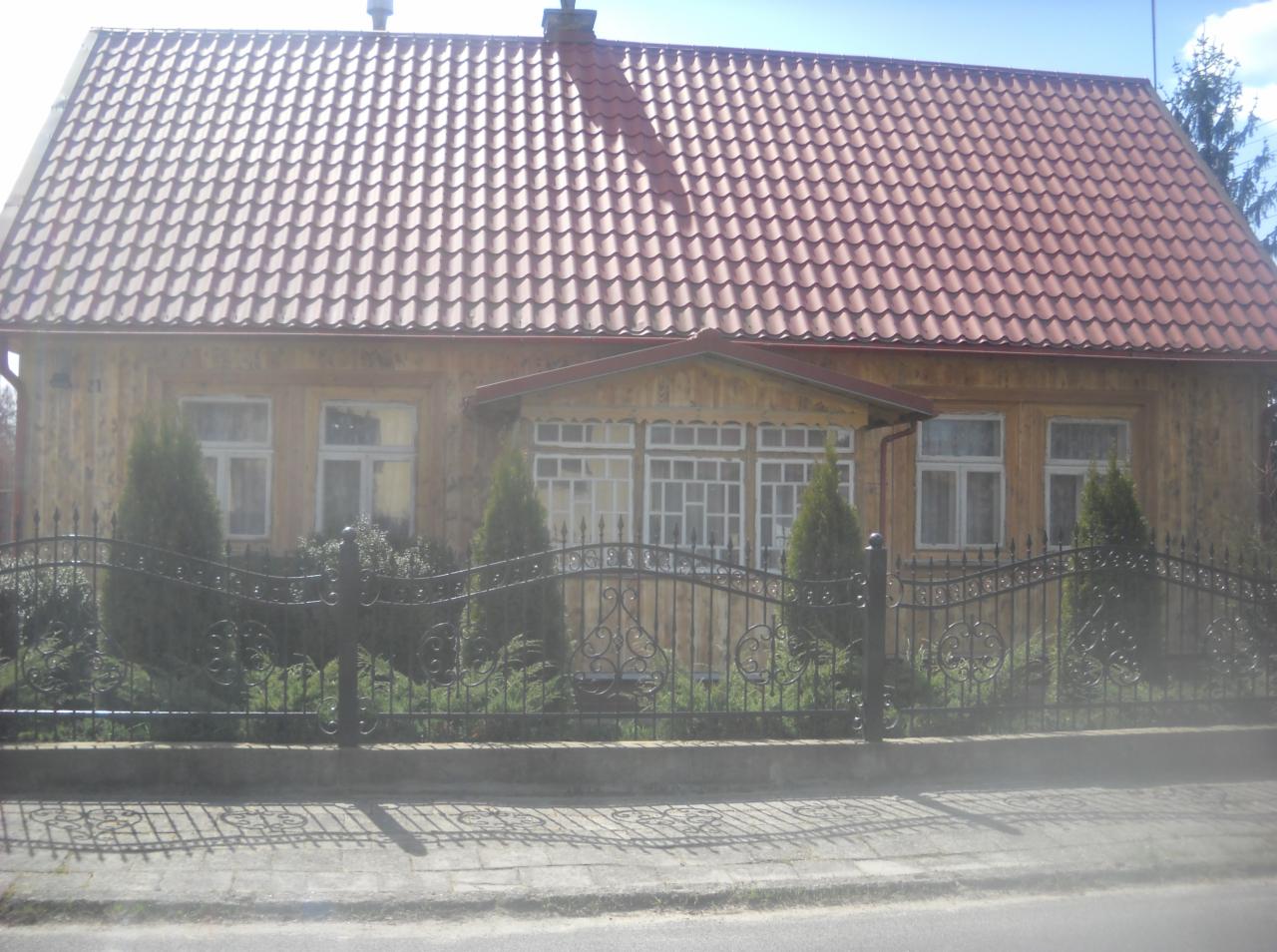 Drewniany dom z czerwonym dachem, ogrodzony kutym, pomalowanym na czarno płotem. Widoczne krzewy i drzewa iglaste. Słoneczny dzień.