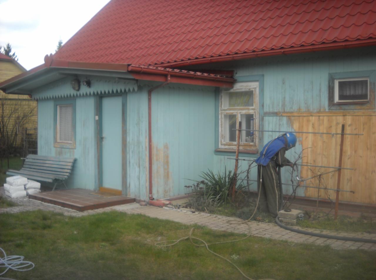 Pracownik w kombinezonie ochronnym czyści metalową konstrukcję przy domu z niebieską elewacją i czerwoną dachówką. Widoczne efekty czyszczenia.