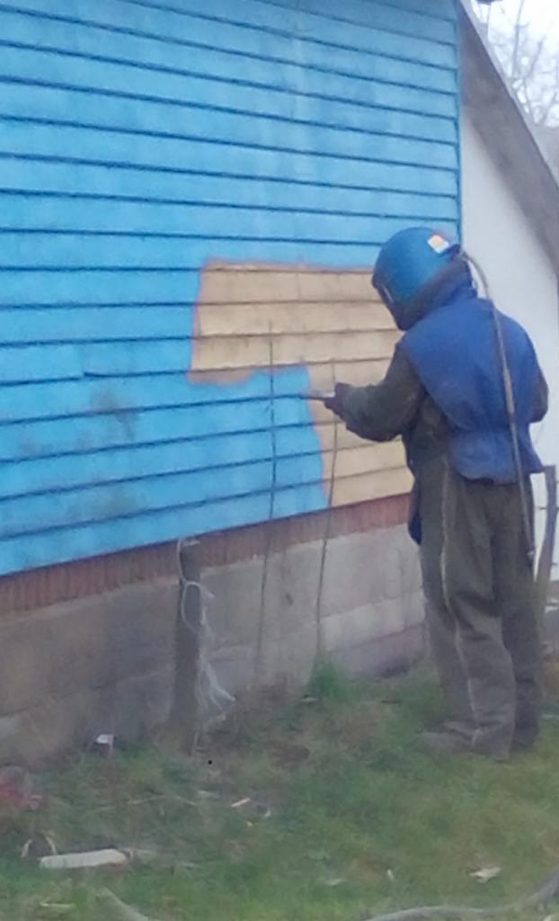 Pracownik w Krosno zabezpiecza elewację drewnianego domu, nakładając farbę ochronną na deski. Widoczny fragment ściany w kolorze błękitnym i surowe drewno.