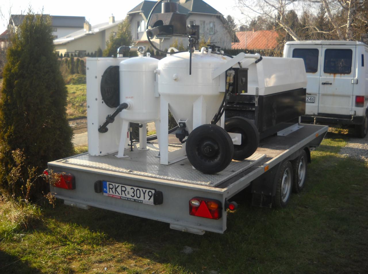 Mobilny zestaw do obróbki powierzchni na przyczepie z widocznym numerem rejestracyjnym, zaparkowany na trawie, w tle domy i biały bus.