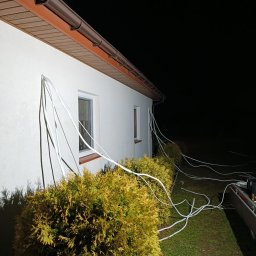 Now-instal - Montaż instalacji klimatyzacji na zewnątrz budynku w Płońsku. Białe przewody prowadzone po elewacji, widoczne krzewy i fragment dachu. Prace w toku, nocne oświetlenie.