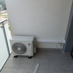 Now-instal - Zamontowana jednostka zewnętrzna klimatyzacji Gree na balkonie z widoczną listwą maskującą przewody. Widok z góry na szare płytki balkonu i fragment ulicy w tle.