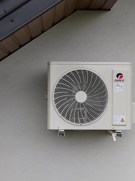 Zamontowana, zewnętrzna jednostka klimatyzacji marki Gree na szarej ścianie budynku. Widoczne mocowania i rura odprowadzająca skropliny. Częściowo widoczny fragment dachu.