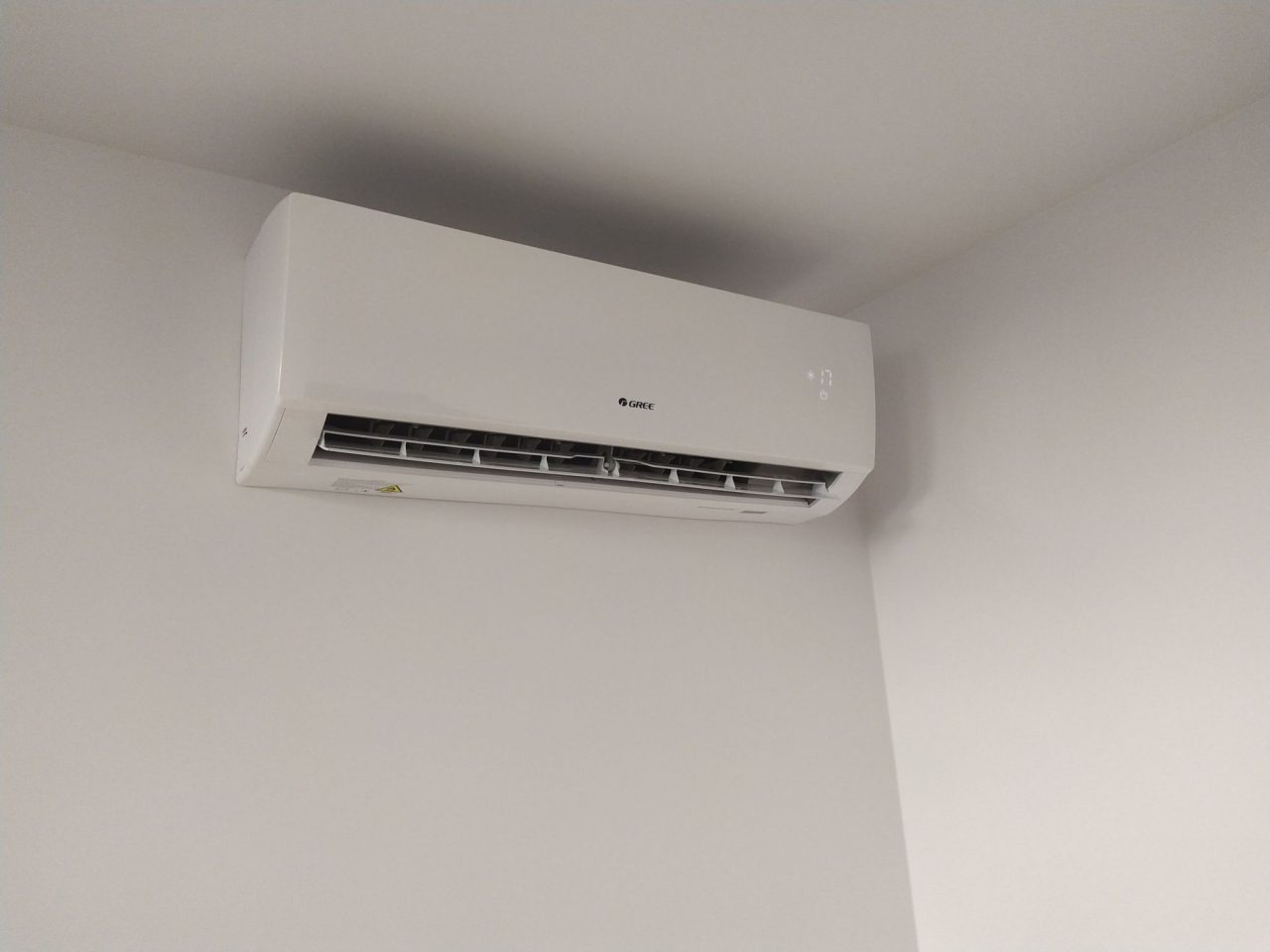 Biała klimatyzacja marki Gree zamontowana na ścianie w rogu pomieszczenia. Widoczny wyświetlacz temperatury i kratka wentylacyjna. Minimalistyczny design.