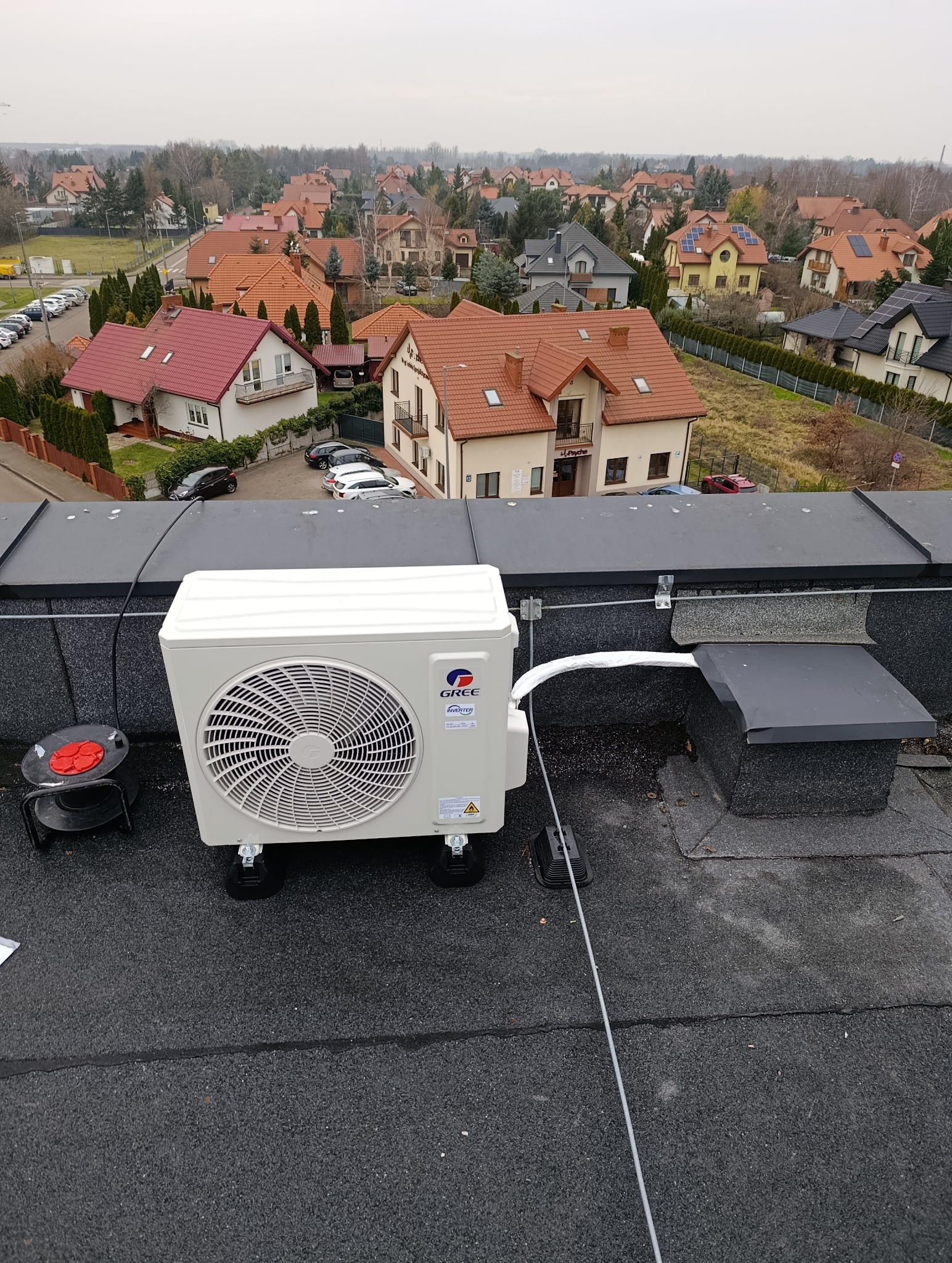 Biały klimatyzator Gree na dachu budynku z widokiem na osiedle domów jednorodzinnych. Widoczne przewody i elementy montażowe.