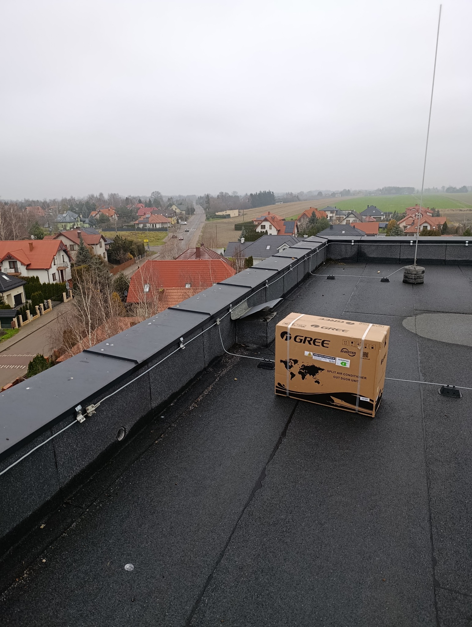 Karton z klimatyzatorem Gree na czarnym dachu budynku z widokiem na zabudowę mieszkalną w tle. Urządzenie gotowe do montażu.