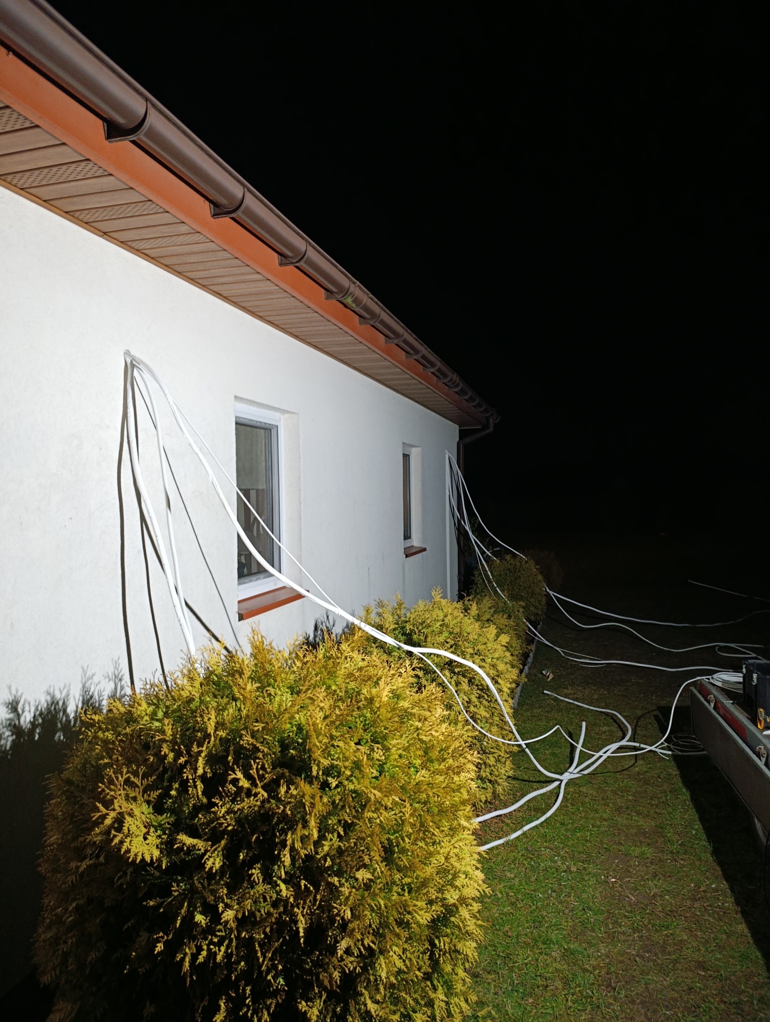 Montaż instalacji klimatyzacji na zewnątrz budynku w Płońsku. Białe przewody prowadzone po elewacji, widoczne krzewy i fragment dachu. Prace w toku, nocne oświetlenie.