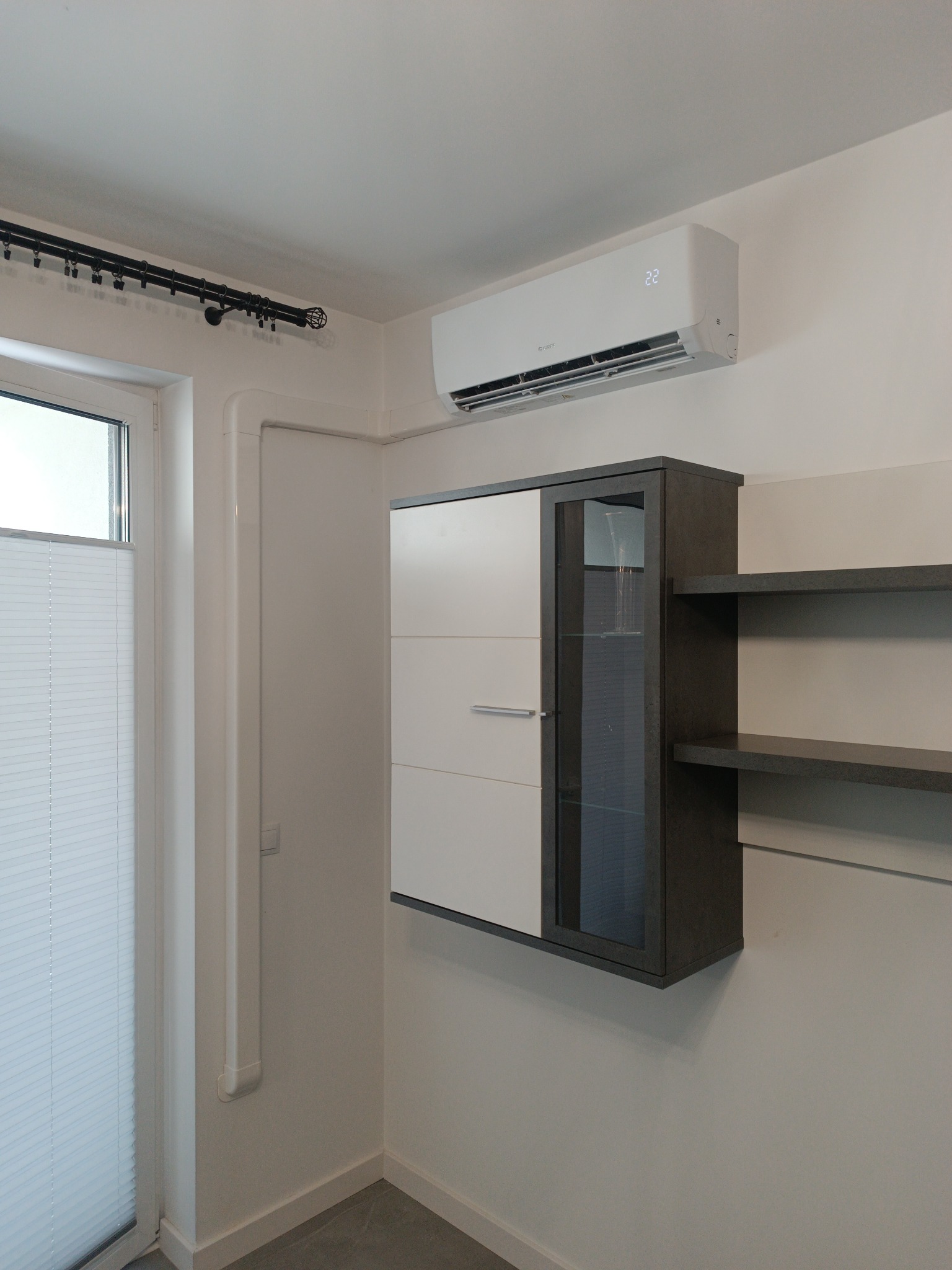 Biały klimatyzator ścienny Gree, zamontowany nad wiszącą szafką z półkami w nowoczesnym wnętrzu. Widoczny fragment okna z żaluzjami i rura instalacyjna.