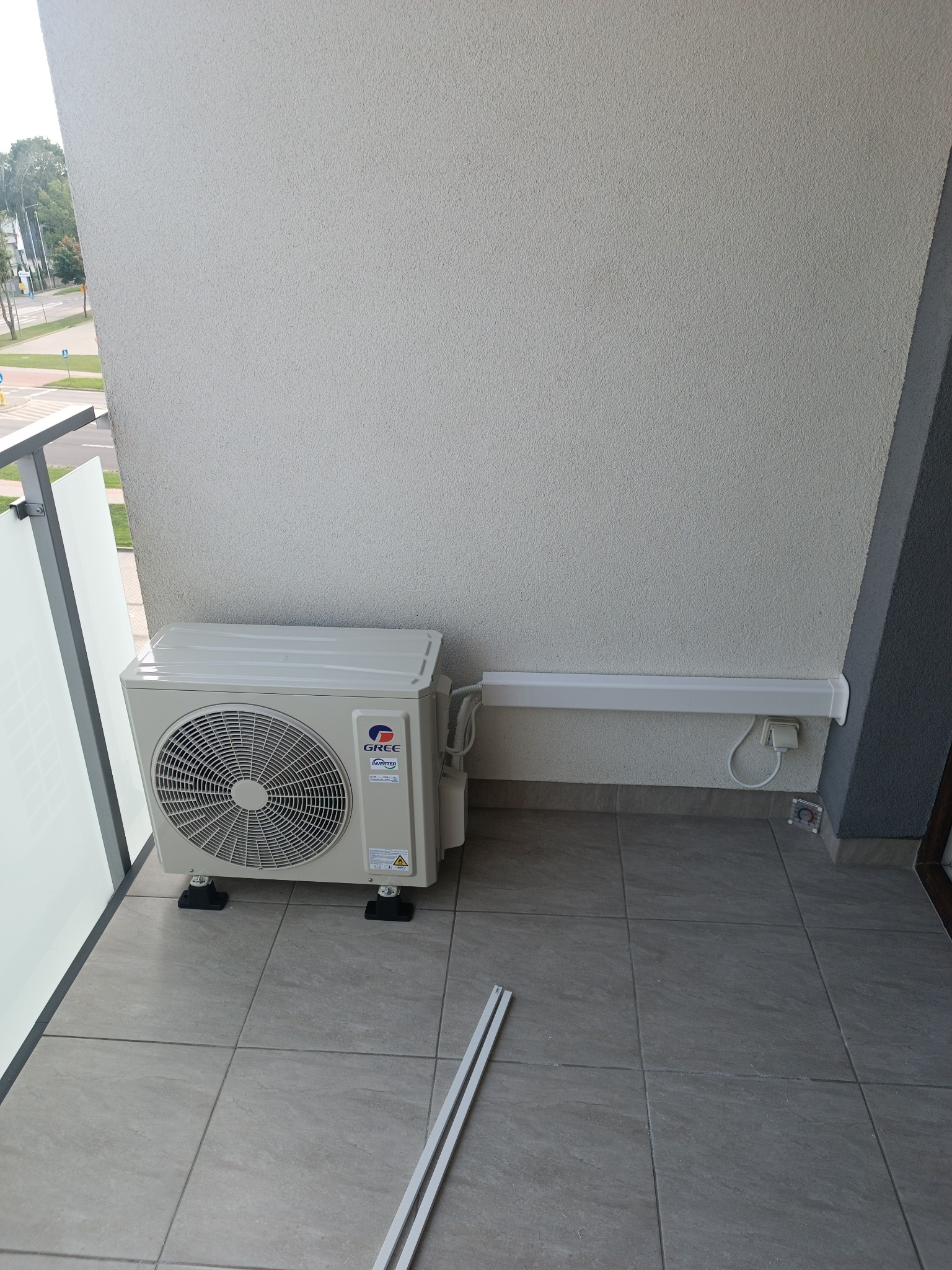 Zamontowana jednostka zewnętrzna klimatyzacji Gree na balkonie z widoczną listwą maskującą przewody. Widok z góry na szare płytki balkonu i fragment ulicy w tle.