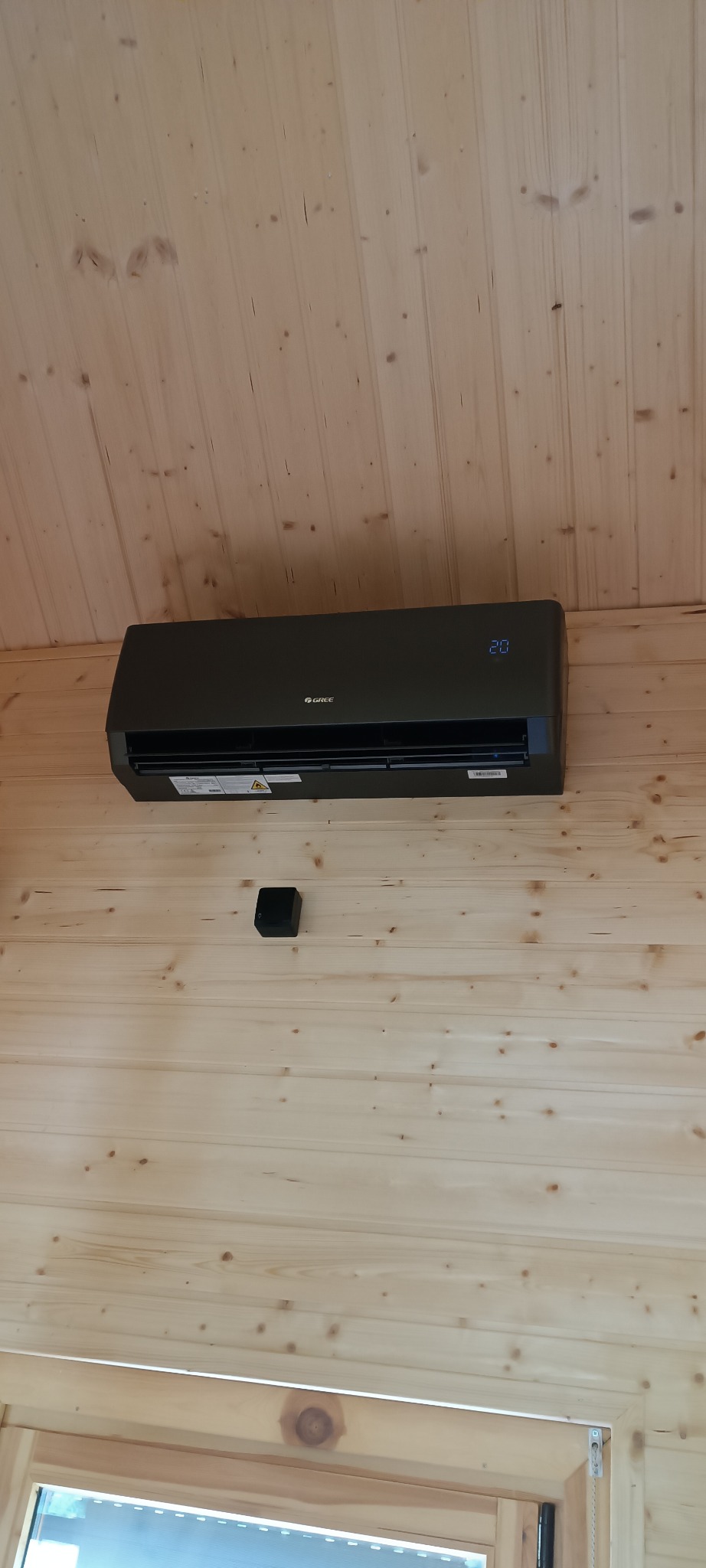 Czarny klimatyzator marki Gree zamontowany na drewnianej ścianie w domu. Widoczny czujnik temperatury. Ustawiona temperatura 20 stopni Celsjusza.