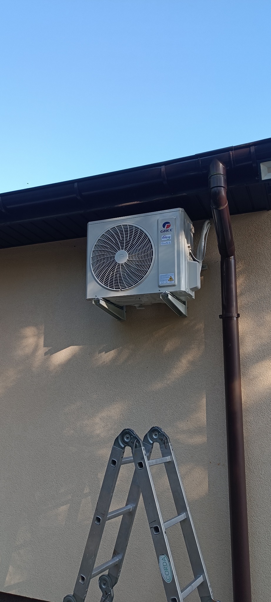 Zamontowana jednostka zewnętrzna klimatyzacji Gree na elewacji budynku, widoczna drabina. Instalacja klimatyzacji w domu jednorodzinnym.