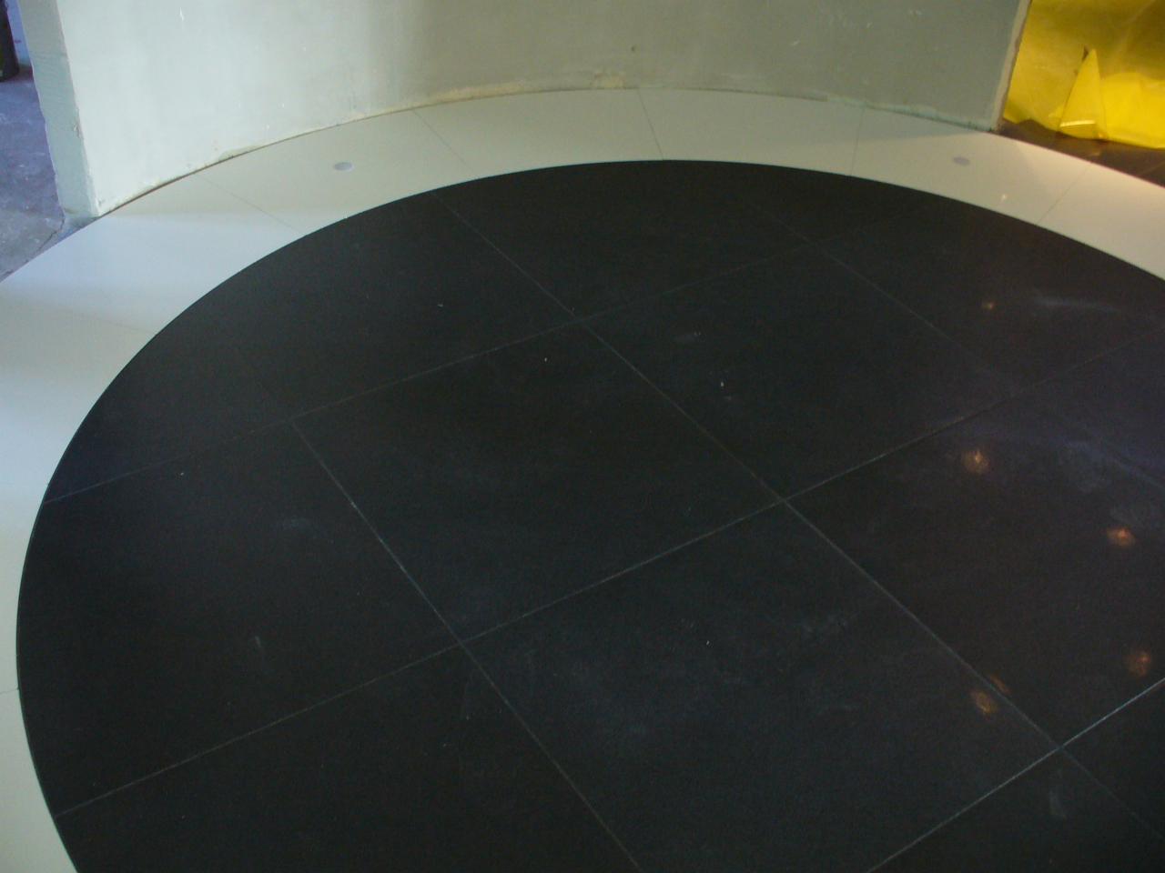 Ciemna, kwadratowa podłoga z płytek ułożona w okrągłym wzorze, otoczona jasnymi płytkami i fragmentem ściany w tle.
