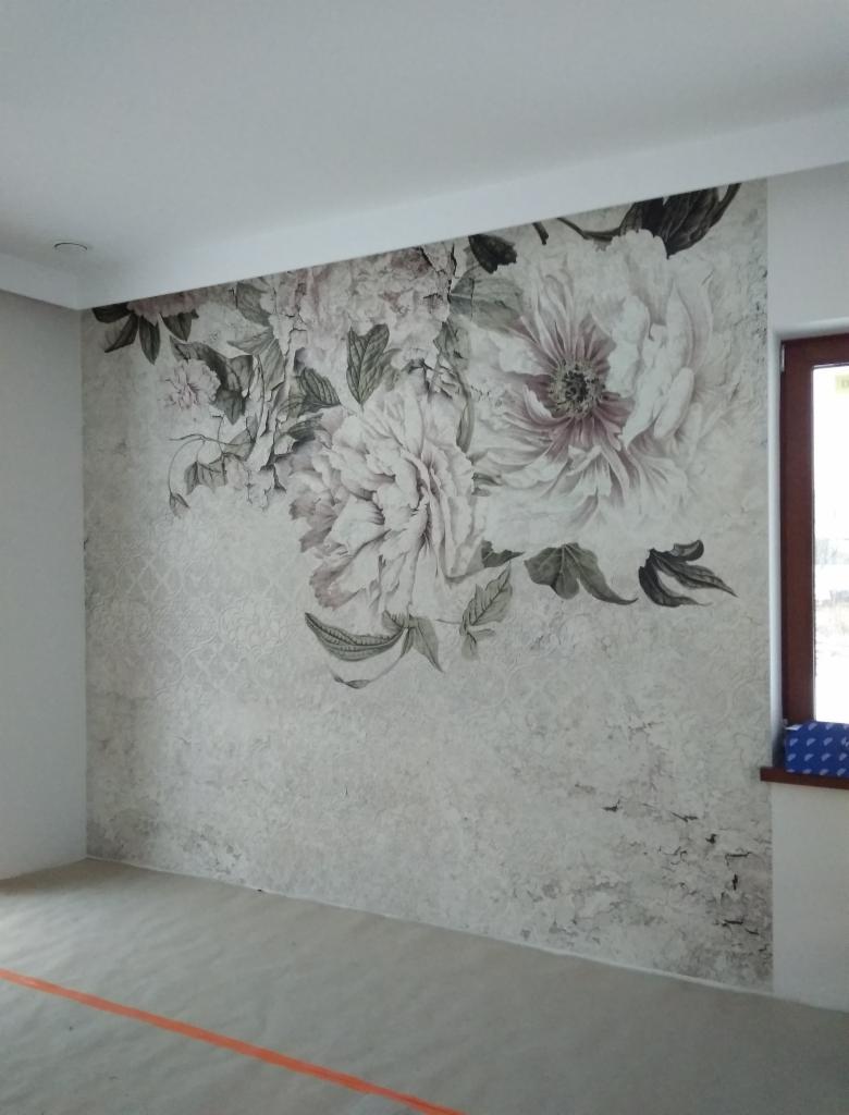 Tapeta z motywem kwiatowym w odcieniach szarości i bieli, na ścianie w pustym pokoju z zabezpieczoną podłogą, widoczna rama okienna.