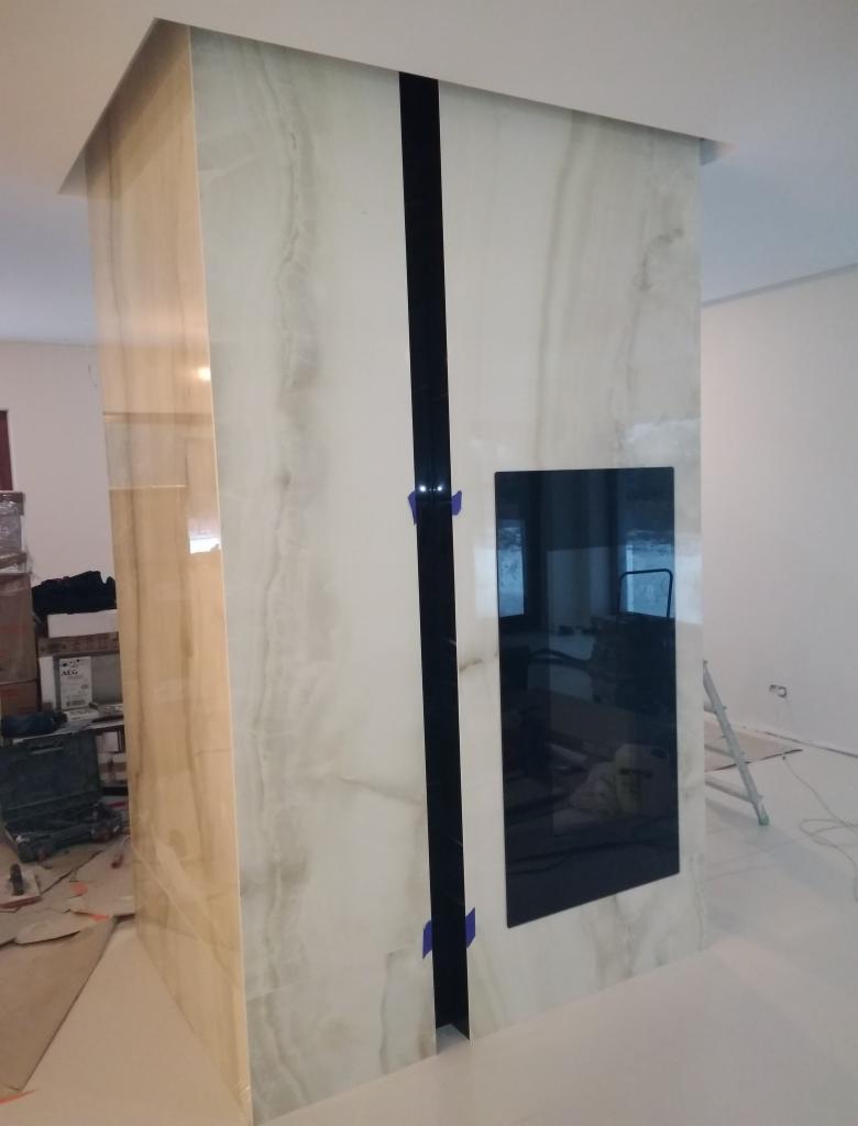 Wnętrze w trakcie prac wykończeniowych: obudowa kominka z płytek imitujących marmur, z czarnym pionowym pasem i wnęką na wkład kominkowy, zabezpieczona niebieską taśmą malarską, widoczne narzędzia...