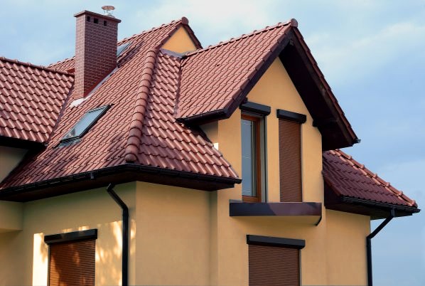 Dom z beżową elewacją i czerwoną dachówką karpiówką, widoczne okna dachowe i brązowe rolety zewnętrzne. Ujęcie z dołu, fragment budynku.