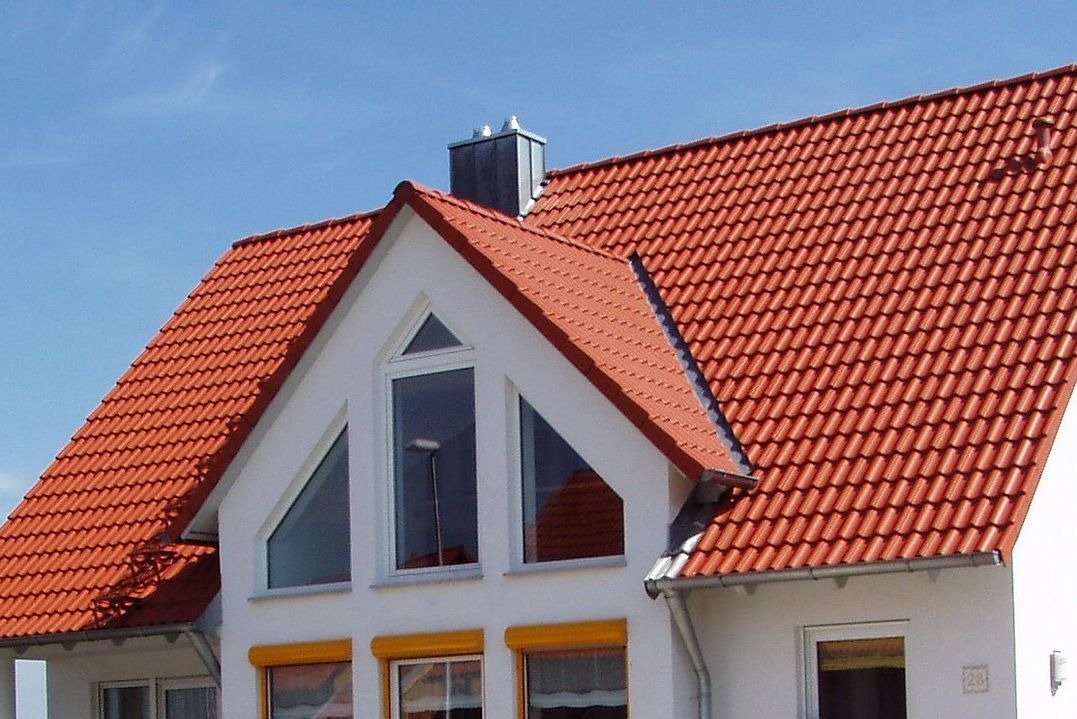 Czerwona dachówka na białym domu z oknami o nietypowym kształcie. Widoczny komin i rynna. Błękitne niebo w tle.