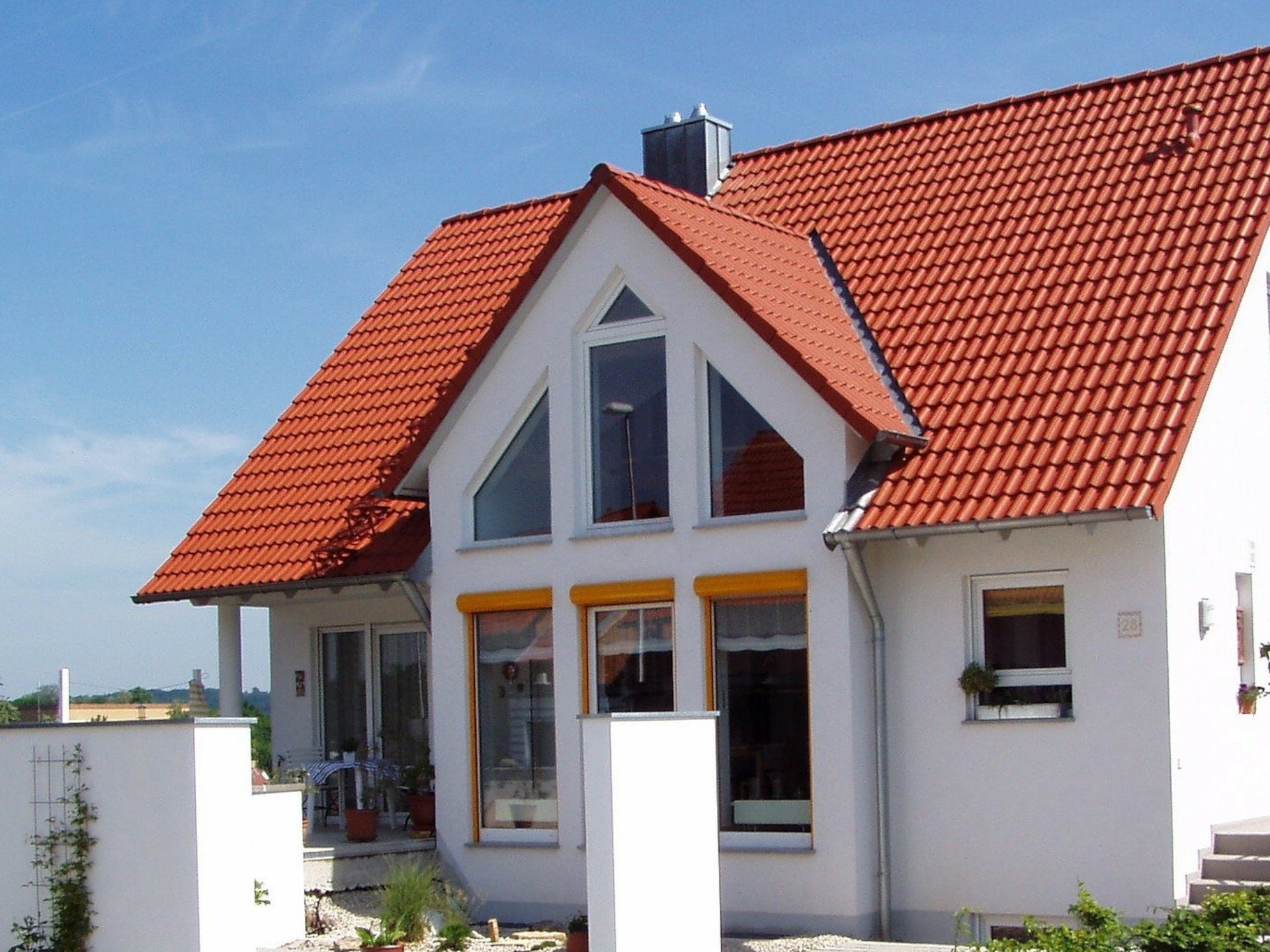 Biały dom jednorodzinny z czerwonym dachem z dachówki ceramicznej, widok z zewnątrz w słoneczny dzień. Żółte rolety okienne.