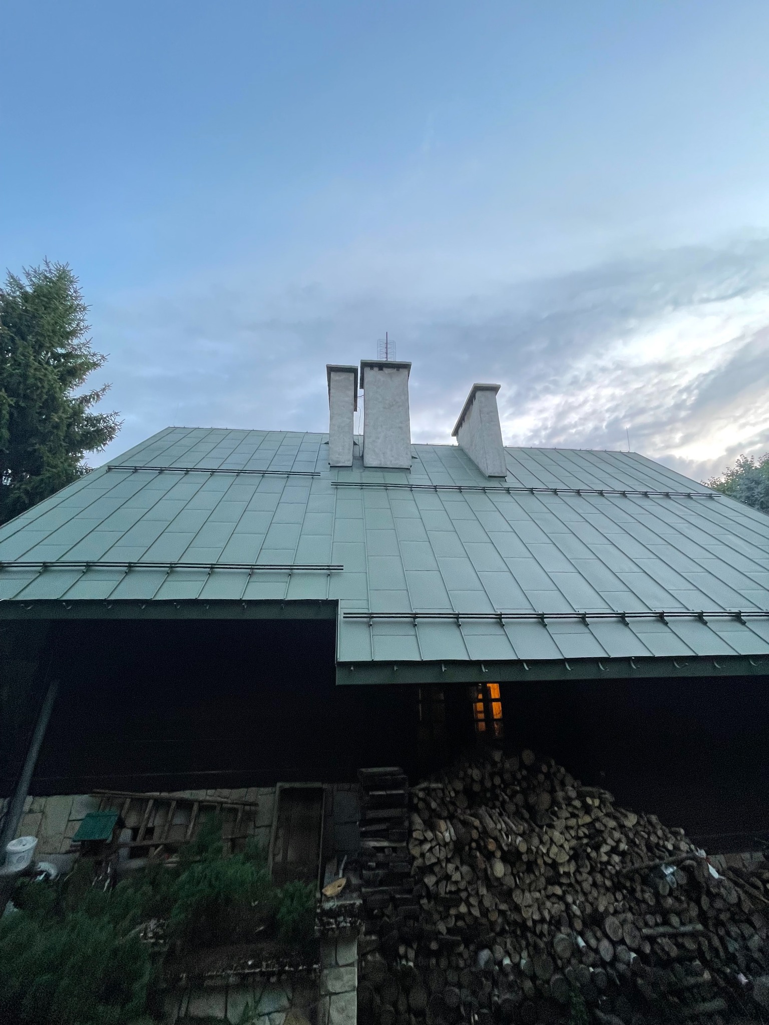 Zielony dach z blachy na drewnianym domu z trzema kominami, widok z dołu na tle pochmurnego nieba. Stos drewna opałowego przy ścianie.