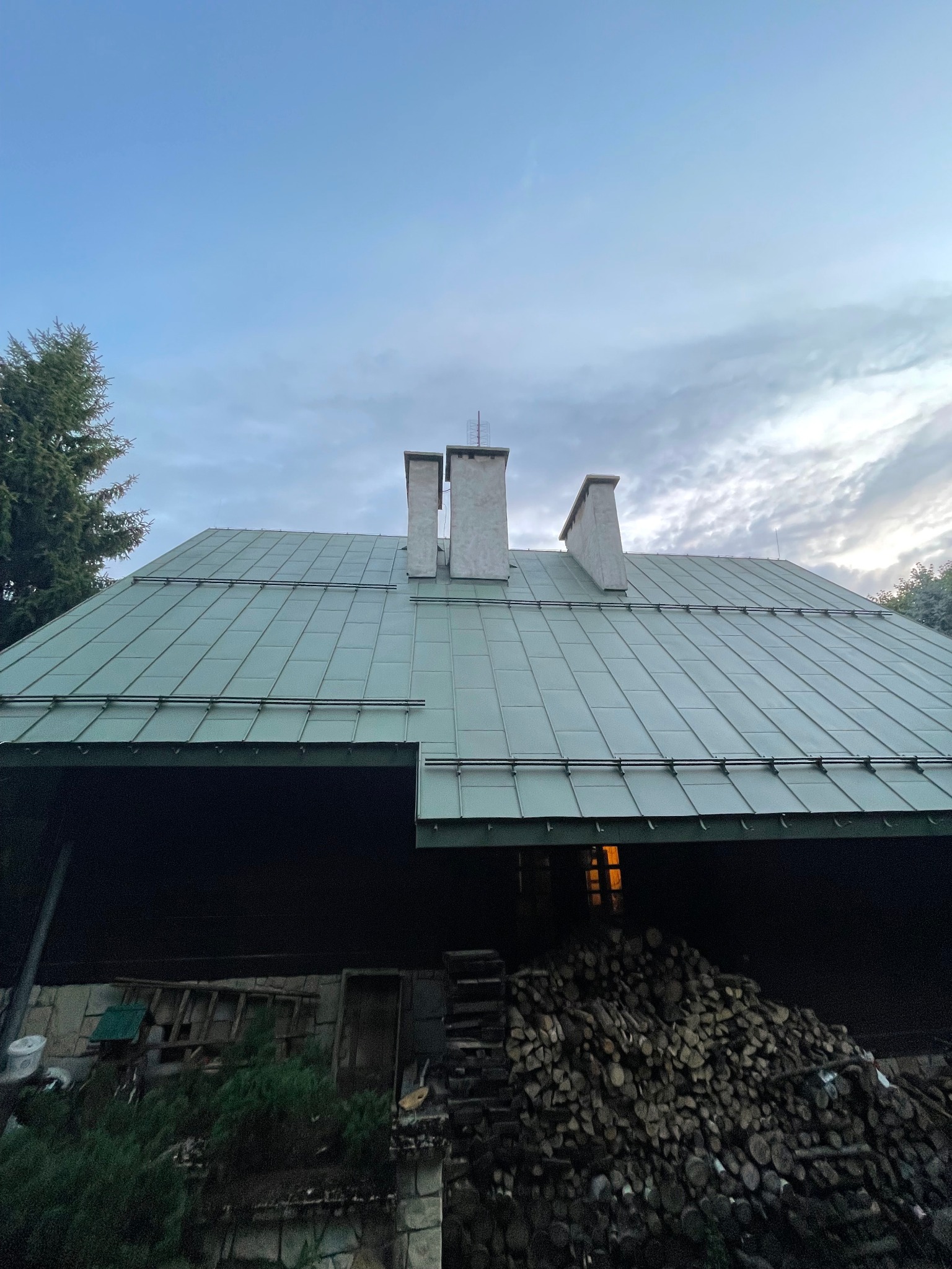Zielony dach z blachy na drewnianym domu z trzema kominami, na tle pochmurnego nieba o zmierzchu. Stos drewna pod okapem. Widoczna drabina i zieleń.