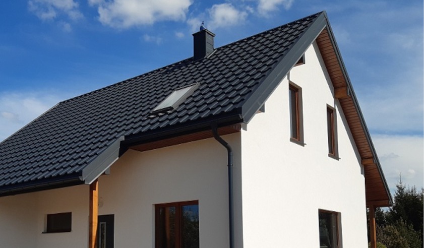 Dom jednorodzinny z ciemnoszarym dachem z dachówki, białymi ścianami i brązowymi ramami okiennymi na tle błękitnego nieba z chmurami. Widoczny fragment rynny.