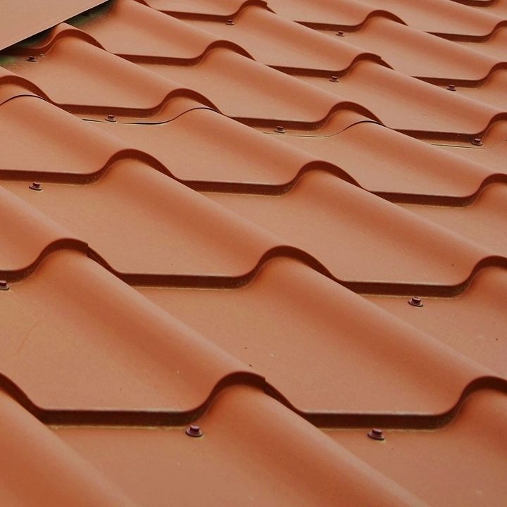 Czerwona dachówka ceramiczna w zbliżeniu, ułożona w rzędach, widoczne mocowania. Regularny wzór, ciepłe oświetlenie, fragment dachu w górnej części kadru.