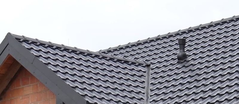 Szary dach z dachówki ceramicznej, widoczny komin wentylacyjny i fragment muru z cegły. Ujęcie z dołu, podkreślające strukturę i geometrię pokrycia dachowego.