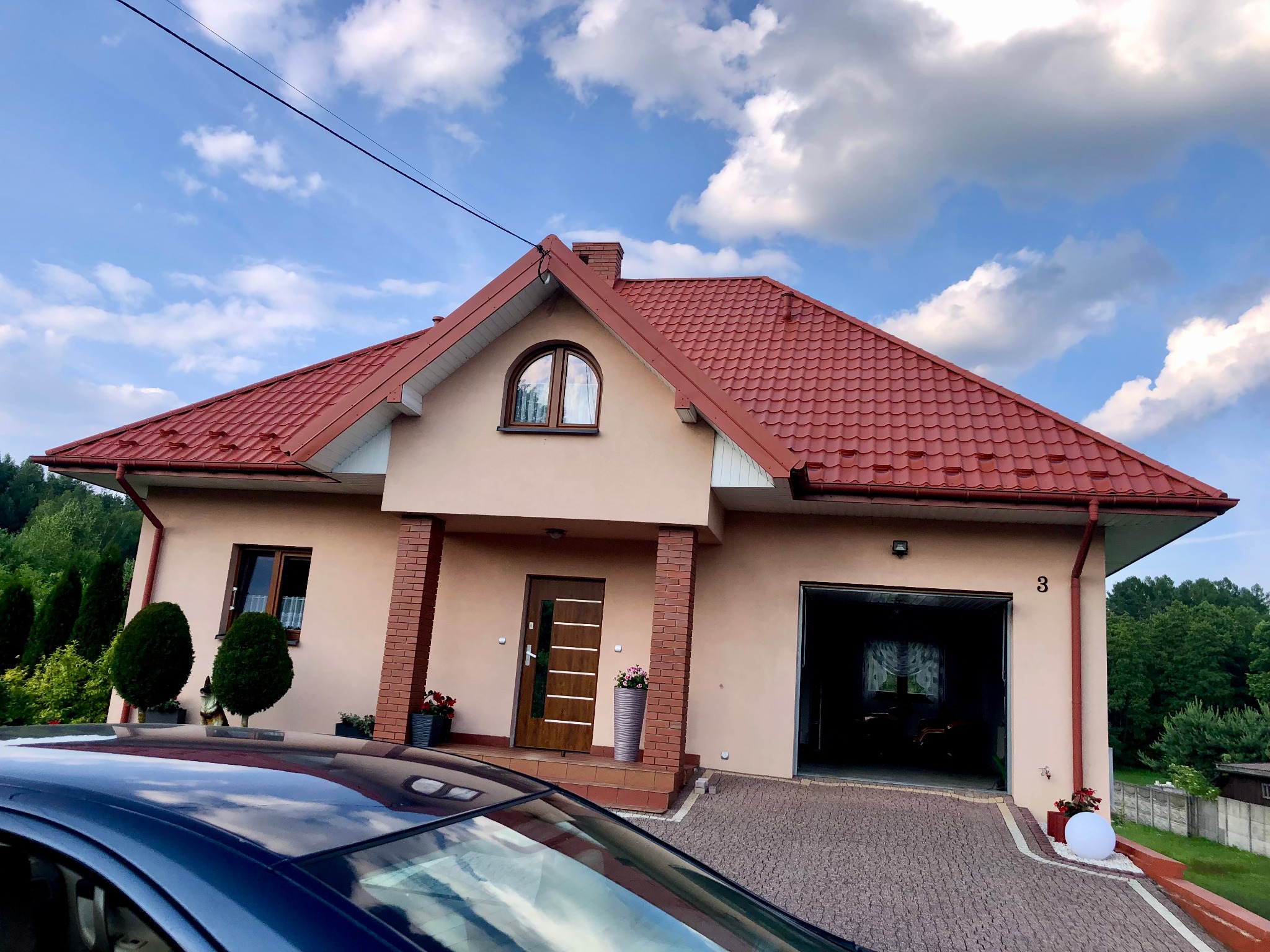 Dom jednorodzinny z czerwoną dachówką, beżową elewacją i otwartym garażem. Na pierwszym planie fragment samochodu. Błękitne niebo z chmurami w tle.
