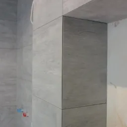 Wykończona ściana w łazience z szarymi, matowymi płytkami imitującymi kamień. Widoczne fugi i narożnik obłożony płytkami. Minimalistyczny design, surowy klimat.