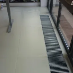 Jasne płytki podłogowe z liniowym odpływem pod oknem, widoczne detale wykończenia i subtelne światło naturalne. Minimalistyczny design wnętrza.