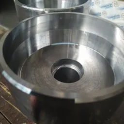 Dwa metalowe, okrągłe elementy z otworem w środku, prawdopodobnie wykonane na tokarce, leżące na drewnianym stole. Widoczne ślady obr&oacute;bki.