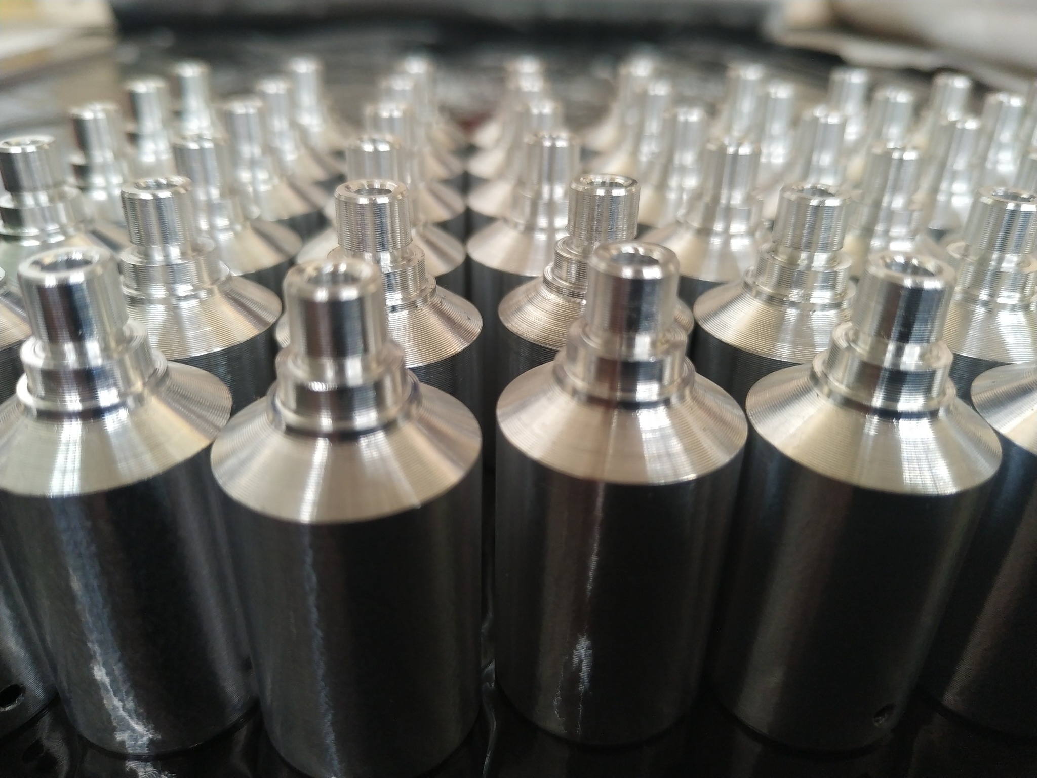 Rząd precyzyjnie obrobionych, srebrnych elementów metalowych po obróbce CNC, ułożonych na czarnej powierzchni. Widoczne ślady obróbki skrawaniem.