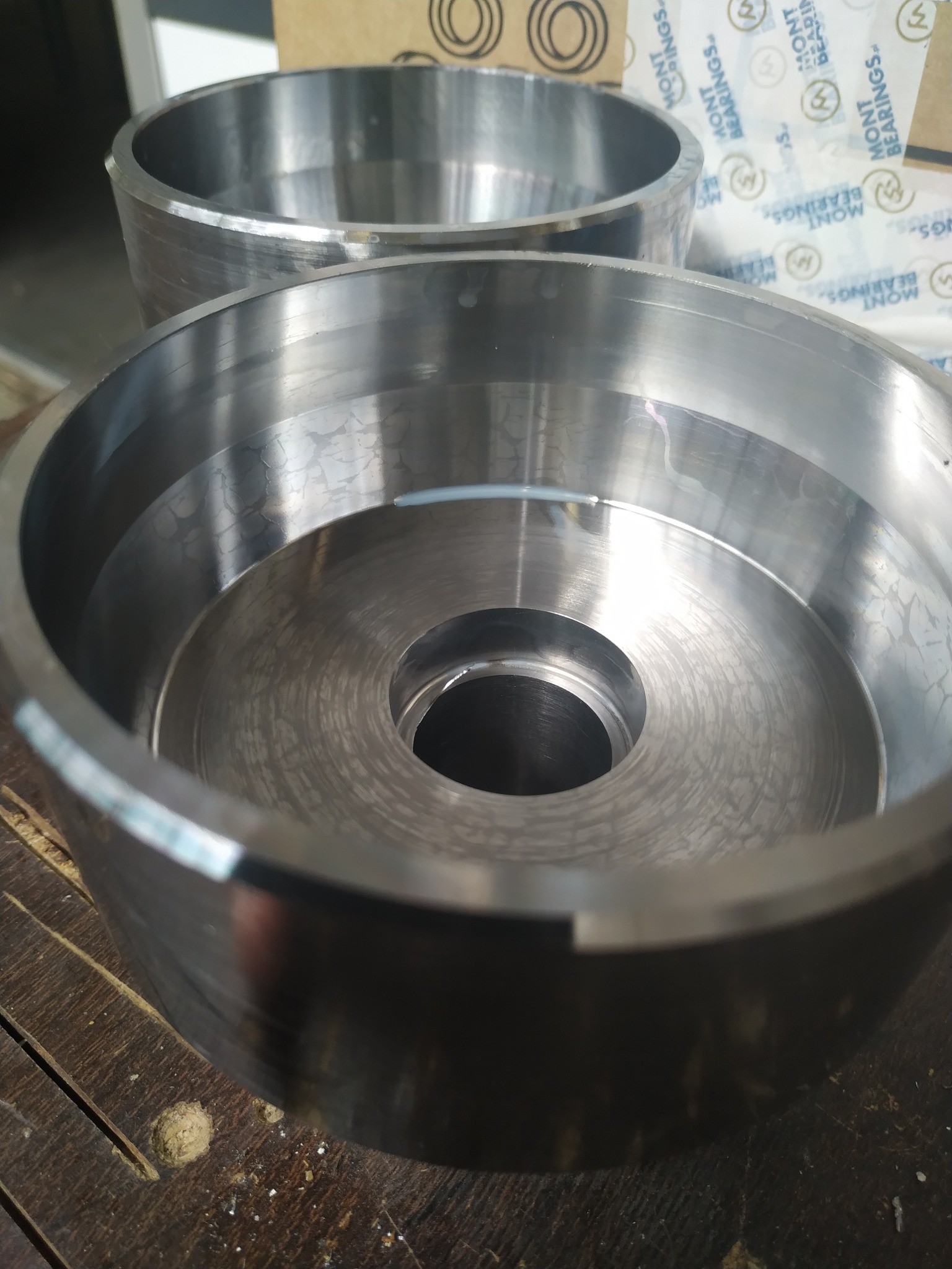 Dwa metalowe, okrągłe elementy z otworem w środku, prawdopodobnie wykonane na tokarce, leżące na drewnianym stole. Widoczne ślady obróbki.