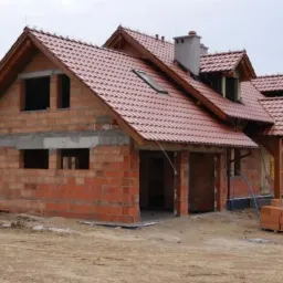 Dom w trakcie budowy z częściowo ukończonym dachem z czerwonej dachówki, widoczna więźba dachowa i murowane ściany z pustaków ceramicznych.