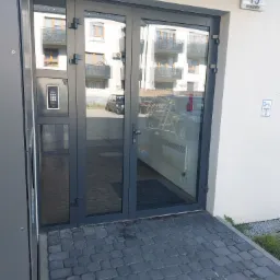 Szklane drzwi wejściowe do budynku z widocznym numerem 15 na ścianie, odbijające otoczenie, w tym samochody i budynek mieszkalny.