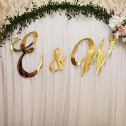 Dekoracja ślubna z białej tkaniny, kwiatów i zieleni, z błyszczącymi złotymi literami E & W na tle wzorzystej ściany.
