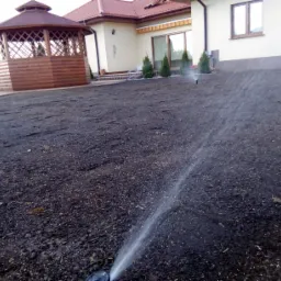 Automatyczny system nawadniania w akcji na świeżo przygotowanym podłożu pod trawnik, w tle altana ogrodowa i dom jednorodzinny.