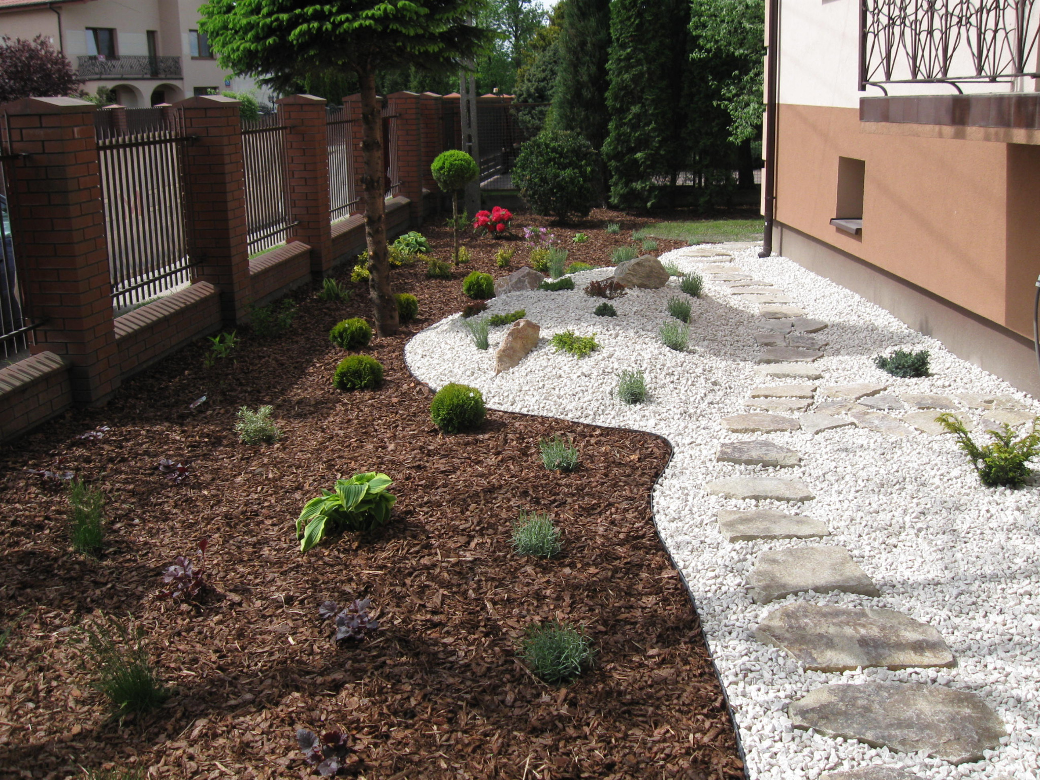 Ogród po realizacji: kora, białe kamienie, ścieżka z płyt kamiennych, ozdobne krzewy i drzewa. Widok na budynek i ogrodzenie z metalowymi przęsłami.