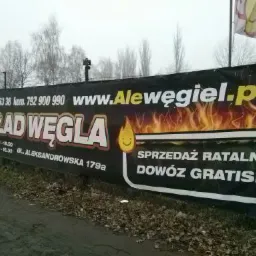 Baner reklamowy składu węgla z adresem www.Alewegiel.pl, numerami telefonów i informacją o sprzedaży ratalnej oraz darmowym dowozie. Widoczne płomienie i symbol kropli.
