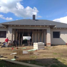 Specjalisci Od Klinkieru - Budowa domu z białej cegły silikatowej w trakcie realizacji, widoczne elementy konstrukcyjne dachu i komina, pracownicy w trakcie pracy, teren budowy z materiałami budowlanymi.