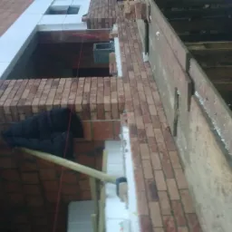 Wznoszenie ściany z cegły klinkierowej z widocznym ociepleniem styropianem, podtrzymywanym drewnianym stemplem i elementami rusztowania.