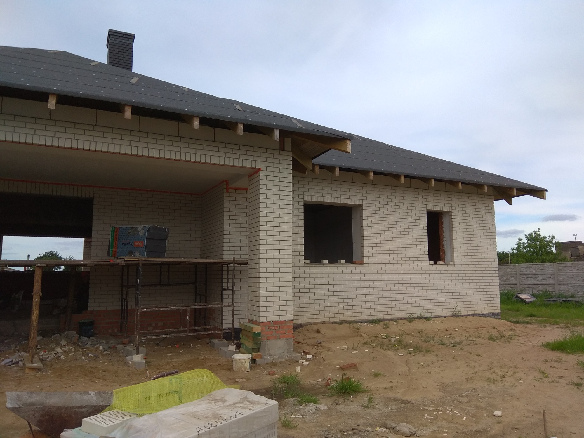 Niedokończony dom murowany z białej cegły silikatowej, widoczne rusztowanie, niezadaszony taras i fragment dachu z kominem, teren wokół w stanie budowy.