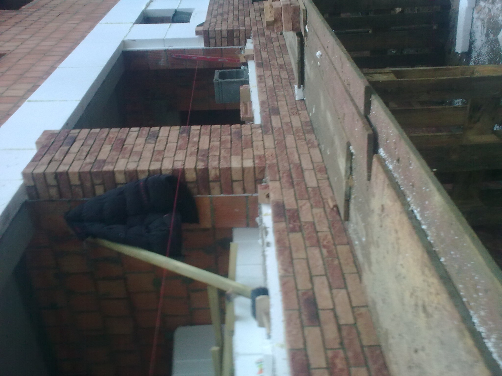 Wznoszenie ściany z cegły klinkierowej z widocznym ociepleniem styropianem, podtrzymywanym drewnianym stemplem i elementami rusztowania.