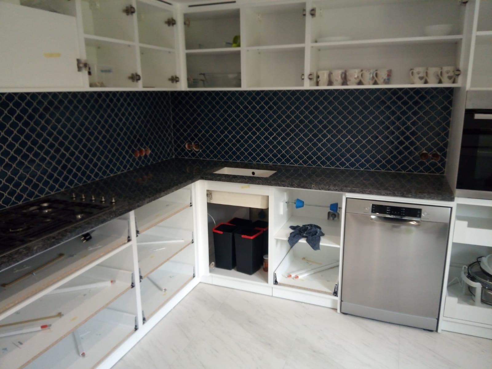 Niebieska mozaika na ścianie nad blatem kuchennym z czarnego granitu, widoczne otwarte szafki bez frontów, srebrna zmywarka w zabudowie i płyta gazowa.